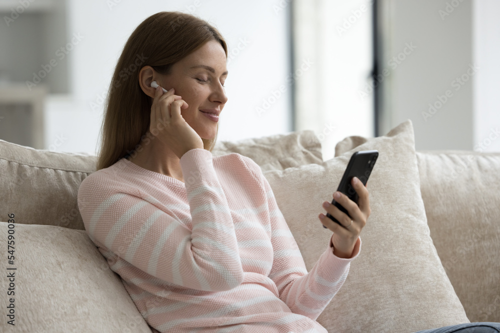 Pretty woman wears wireless earphones play music on cellphone use digital streaming on-line services enjoy favourite song with eyes closed relax on sofa at home. Hobby, stress relief, weekend leisure Pretty woman wears wireless earphones play music on cellphone use digital streaming on-line services enjoy favourite song with eyes closed relax on sofa at home. Hobby, stress relief, weekend leisure