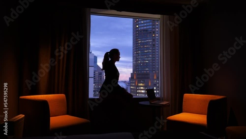 Young woman sitting on window sill in dark room, looking out at the evening city. Blurred background, dark silhouette of pensive lady against the sunset sky. Notebook computer on small table near her