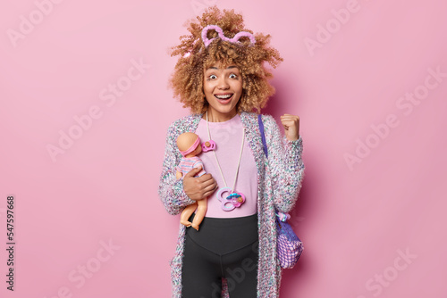 Happy pregnant woman hears great news clenches fist and smiles positively holds doll for future daughter has big tummy looks positively at camera isolated over pink background. Motherhood concept