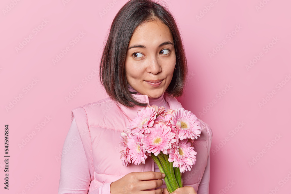 Portrait of pensive young Asian woman with thoughtful expression wears turtleneck and vest holds bouquet of gerbera flowers celebrates birthday isolated over pink background has romantic mood