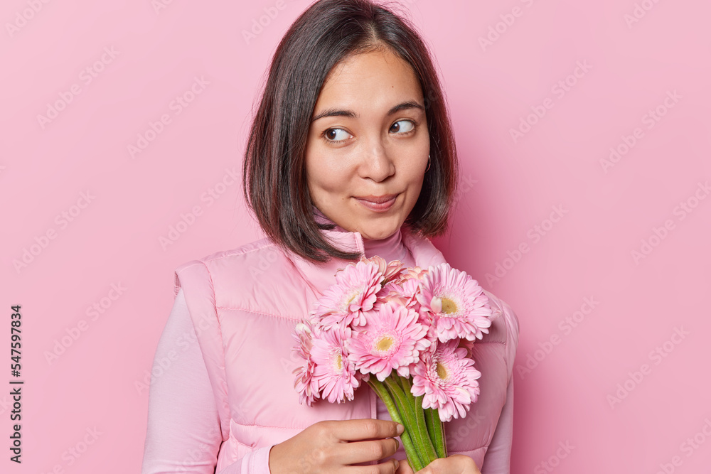 Portrait of pensive young Asian woman with thoughtful expression wears turtleneck and vest holds bouquet of gerbera flowers celebrates birthday isolated over pink background has romantic mood Portrait of pensive young Asian woman with thoughtful expression wears turtleneck and vest holds bouquet of gerbera flowers celebrates birthday isolated over pink background has romantic mood