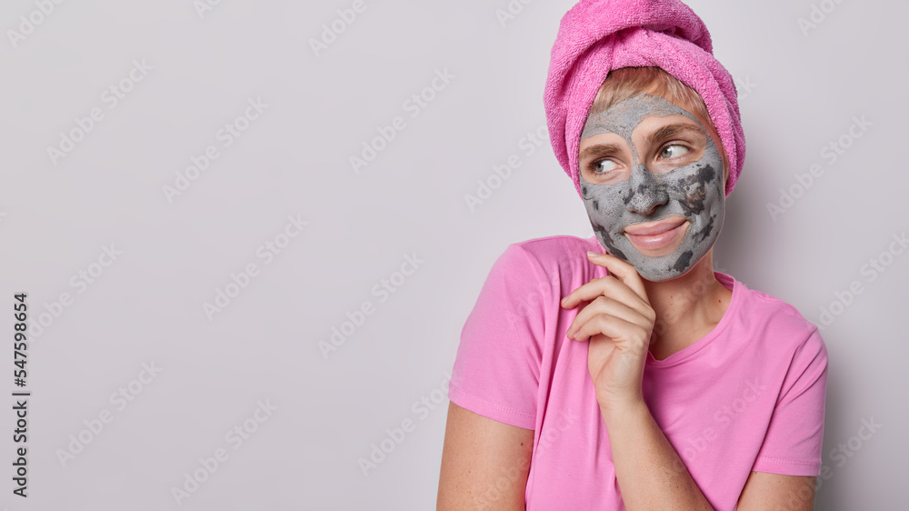 Lovely dreamy woman with big grey eyes undergoes skin care procedures applies facial clay mask touches face gently concentrated aside wears pink t shirt towel on head stands against grey studio wall Lovely dreamy woman with big grey eyes undergoes skin care procedures applies facial clay mask touches face gently concentrated aside wears pink t shirt towel on head stands against grey studio wall