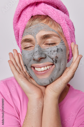 Close up shot of pleased young woman with pink towel on head applies facial clay mask to reduce fine lines blackheads smiles broadly keeps eyes closed undergoes beauty procedures after taking shower