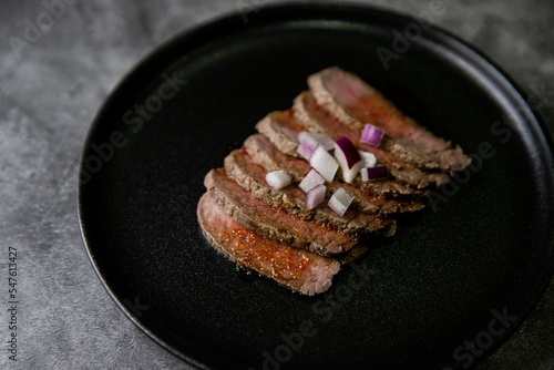 Closeup shot of steak tataki in sauce and onions on black plate with grey blurred background