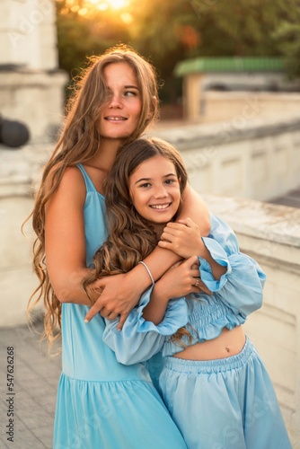 Portrait of mother and daughter in blue dresses with flowing long hair against the backdrop of sunset. The woman hugs and presses the girl to her. They are looking at the camera.