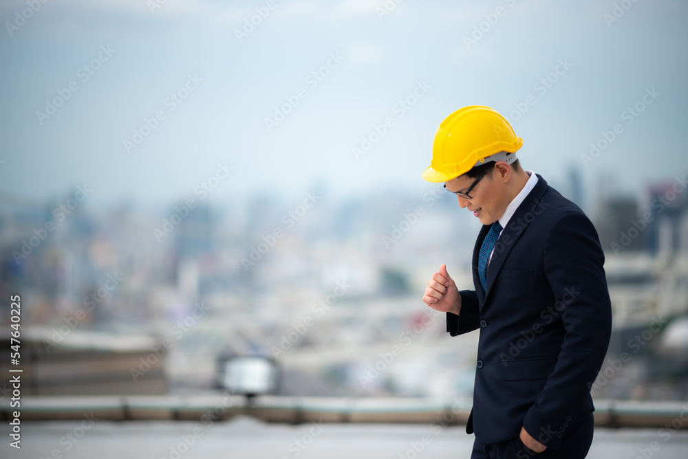 Young business man and architect or contractor in formal clothing with yellow hardhat standing with open arms wearing eyeglasses on rooftop of new building against tall buildings Young business man and architect or contractor in formal clothing with yellow hardhat standing with open arms wearing eyeglasses on rooftop of new building against tall buildings