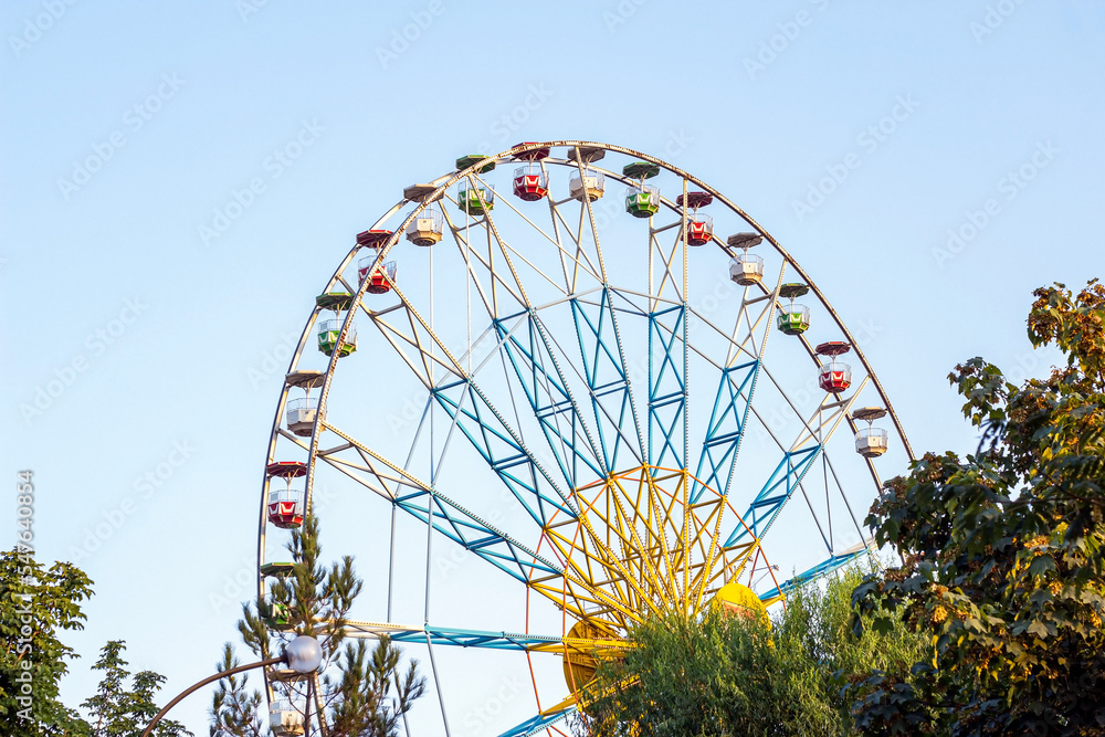 High carnival Ferris wheel in the amusement park in summer spinning against the blue sky. High carnival Ferris wheel in the amusement park in summer spinning against the blue sky.
