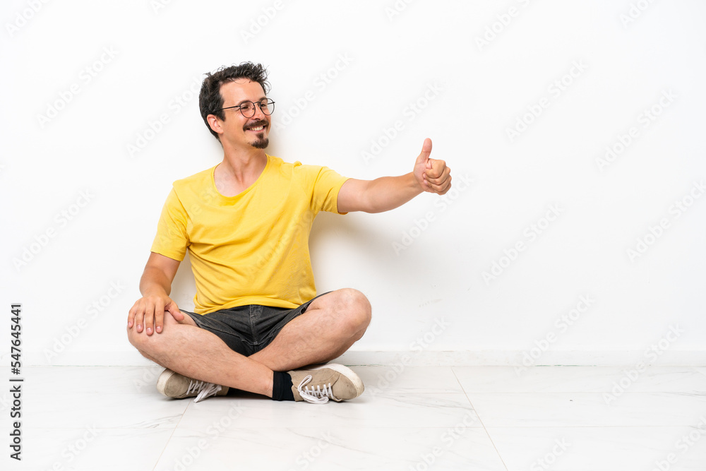Fototapeta premium Young caucasian man sitting on the floor isolated on white background giving a thumbs up gesture