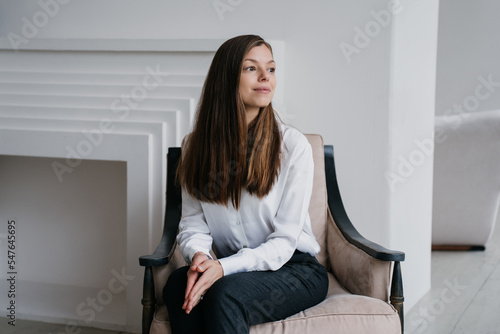 Beautiful Caucasian young woman in white shirt and black pants sitting on chair looks aside dreamily thinking about career and life. Purposeful businesswoman smiling, planing day. Successful people.