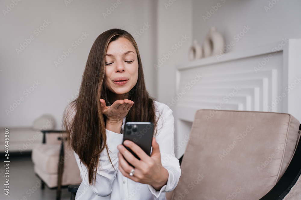 Cheerful Hispanic young woman holds phone sends air kiss during video call sitting on cozy chair at home dressed in white shirt. Pretty brunette businesswoman grateful to her husband. Happy woman. Cheerful Hispanic young woman holds phone sends air kiss during video call sitting on cozy chair at home dressed in white shirt. Pretty brunette businesswoman grateful to her husband. Happy woman.