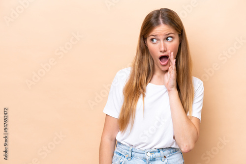 Young caucasian woman isolated on beige background whispering something with surprise gesture while looking to the side