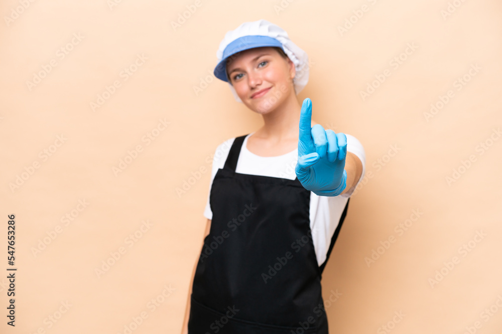 Fishmonger caucasian woman wearing an apron and holding a raw fish isolated on beige background showing and lifting a finger Fishmonger caucasian woman wearing an apron and holding a raw fish isolated on beige background showing and lifting a finger