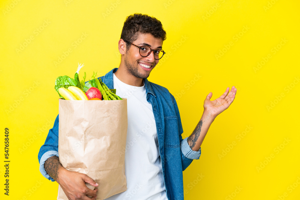Young Brazilian man holding a grocery shopping bag isolated on yellow background extending hands to the side for inviting to come Young Brazilian man holding a grocery shopping bag isolated on yellow background extending hands to the side for inviting to come