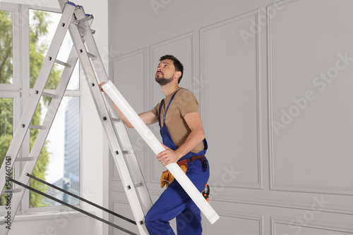 Electrician in uniform with ceiling lamp on metal ladder indoors