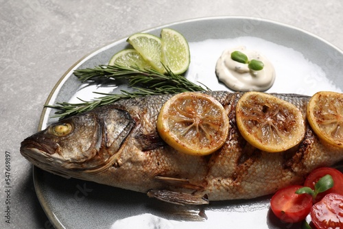 Delicious roasted sea bass fish served with lemon, rosemary and sauce on light grey table, closeup