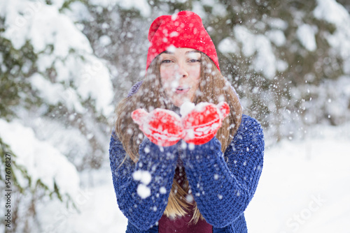 Happy woman in snowy winter day outdoor. Female model dressed bright sweater and red hat blowing snow