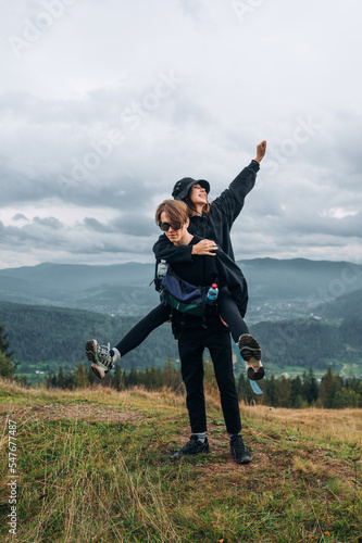 A young happy couple having fun in the mountains during a hike, a man carries a woman on his back with a background of beautiful mountain scenery.