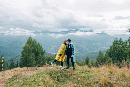 Cute couple of tourists in raincoats standing in the mountains, hugging and kissing during the sunset on the background of cloudy mountain views