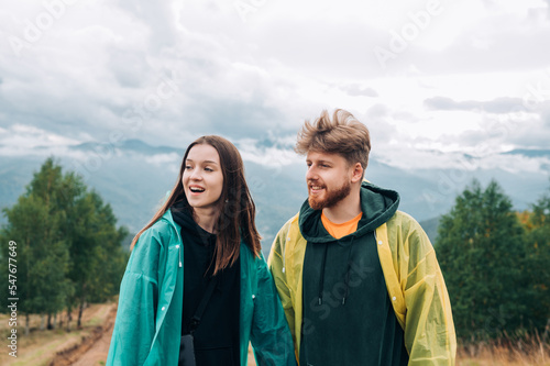 Photo of a happy couple of tourists on a hike standing on the top of a mountain wearing raincoats and looking away with a smile on their face