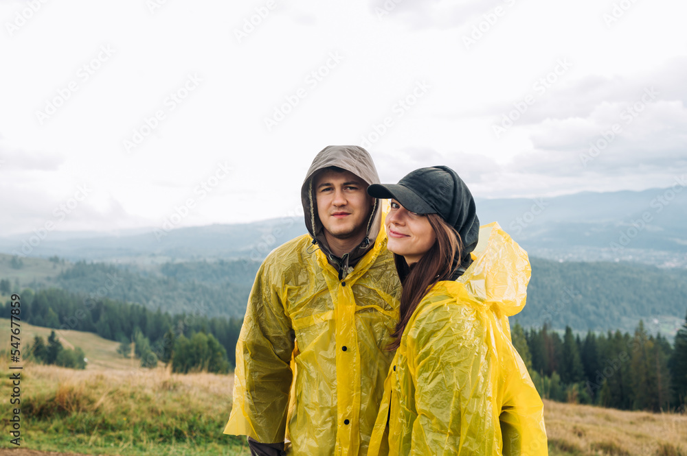 Beautiful young couple man and woman in raincoats standing with smiles on their faces against the background of mountain views and posing for the camera with a serious face.