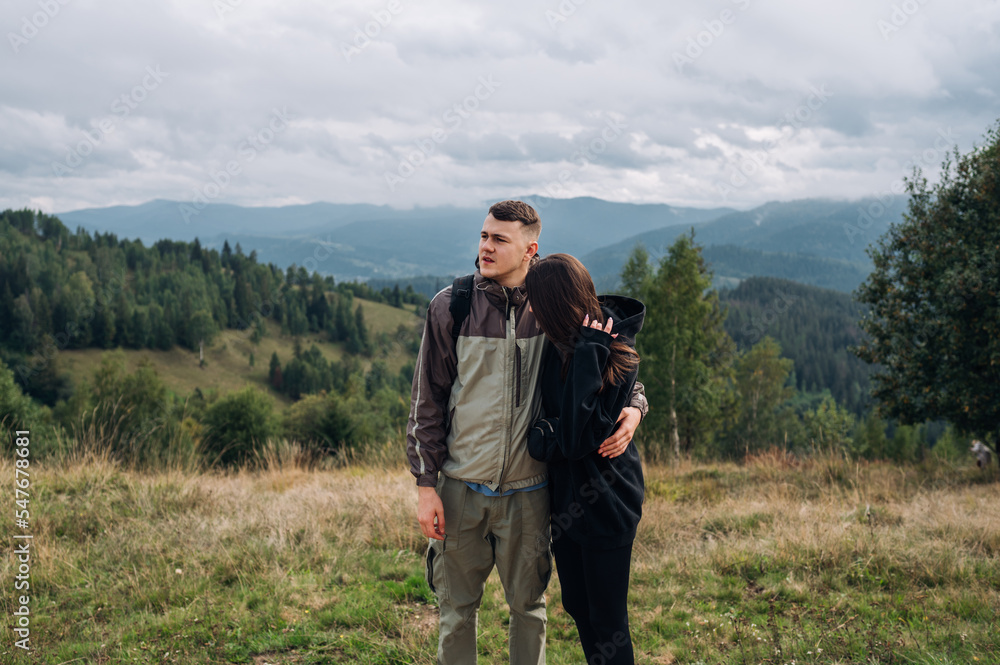 a beautiful couple of tourists standing in the mountains during a hike and hugging each other, looking away on the background of beautiful scenery.