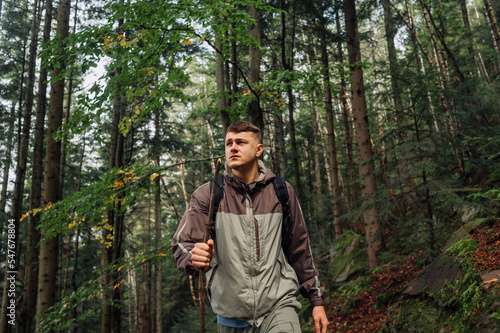A handsome male hiker with a backpack and a stick in his hand is walking in the mountains through a dense forest with a serious face and looking away. Active tourism.м