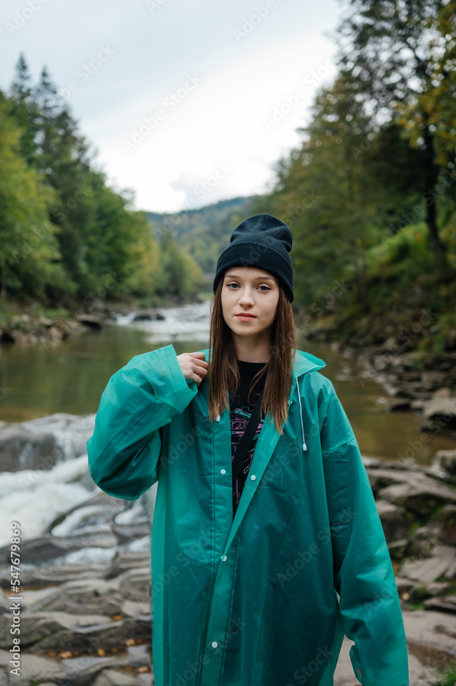 A beautiful tourist girl in casual clothes and a raincoat stands on the background of a mountain river and beautiful views of the forest and looks at the camera with a serious face.