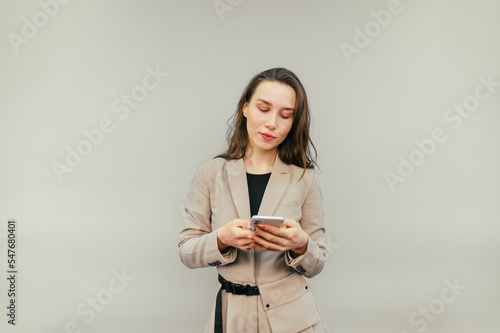 Beautiful woman in smart clothes uses a smartphone on a beige background, looks at the screen with a calm face, wears a beige jacket.