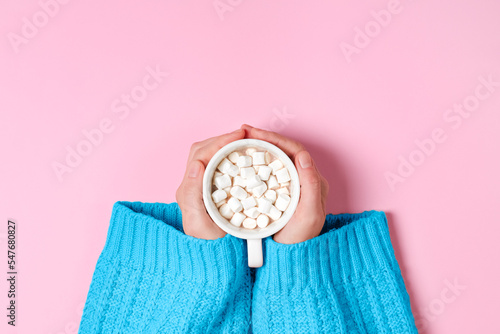 Top view on Girls hands in blue cozy oversized sweater holding large cup of cocoa or hot chocolate isolated on pink background.
