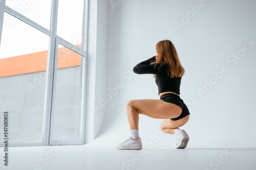 Fototapeta Naklejka Na Ścianę i Meble -  Sexy woman in short shorts with red hair sitting by the window on a white background and dancing twerk