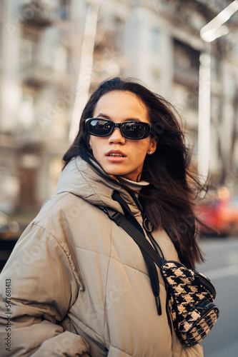 Close-up portrait of a hispanic woman in sunglasses standing outdoors in a warm jacket and looking away with a serious face. © bodnarphoto