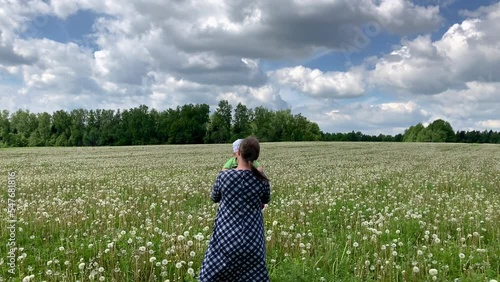 Mom and child are circling on a green field. A young woman plays with a mouse in her arms while tossing and circling the little one. The family walks through a flowering dandelion field.