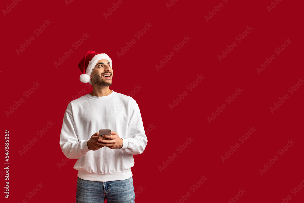 Cheerful Arab Man Wearing Santa Hat Holding Smartphone And Looking Aside Cheerful Arab Man Wearing Santa Hat Holding Smartphone And Looking Aside