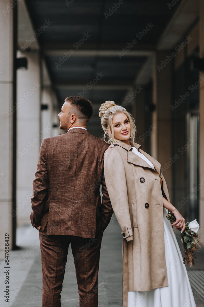 A beautiful blonde model bride with a bouquet of flowers in a beige coat stands in a city tunnel, leaning on a groom in a brown stylish suit. Wedding photography, portrait, autumn.