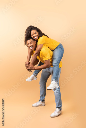 Young black man giving piggyback ride to his girlfriend against peach studio background, full length shot