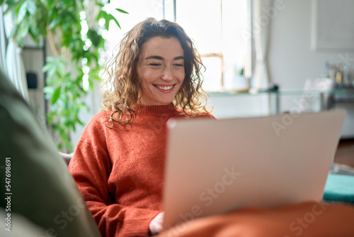 Relaxed happy young woman sitting on sofa using laptop at home surfing, doing online ecommerce shopping, looking at computer ordering sale products on website, watching videos or elearning.