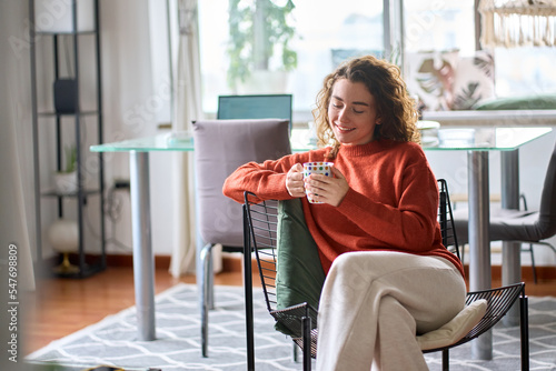 Young adult smiling pretty woman sitting on chair holding cup drinking tea or coffee relaxing at home. Happy calm lady enjoying warm hot drink with mug in hands daydreaming in cold cozy morning.