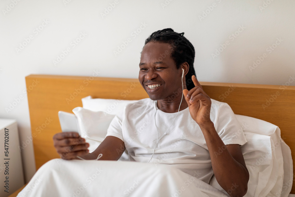 Cheerful Black Man Relaxing In Bed With Smartphone And Earphones Cheerful Black Man Relaxing In Bed With Smartphone And Earphones