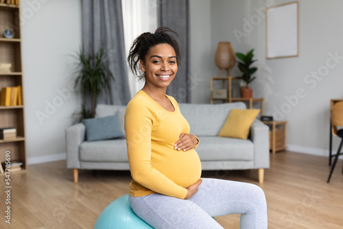 Cheerful young black pregnant lady future mom with big belly do exercises on fit ball, feels baby movements
