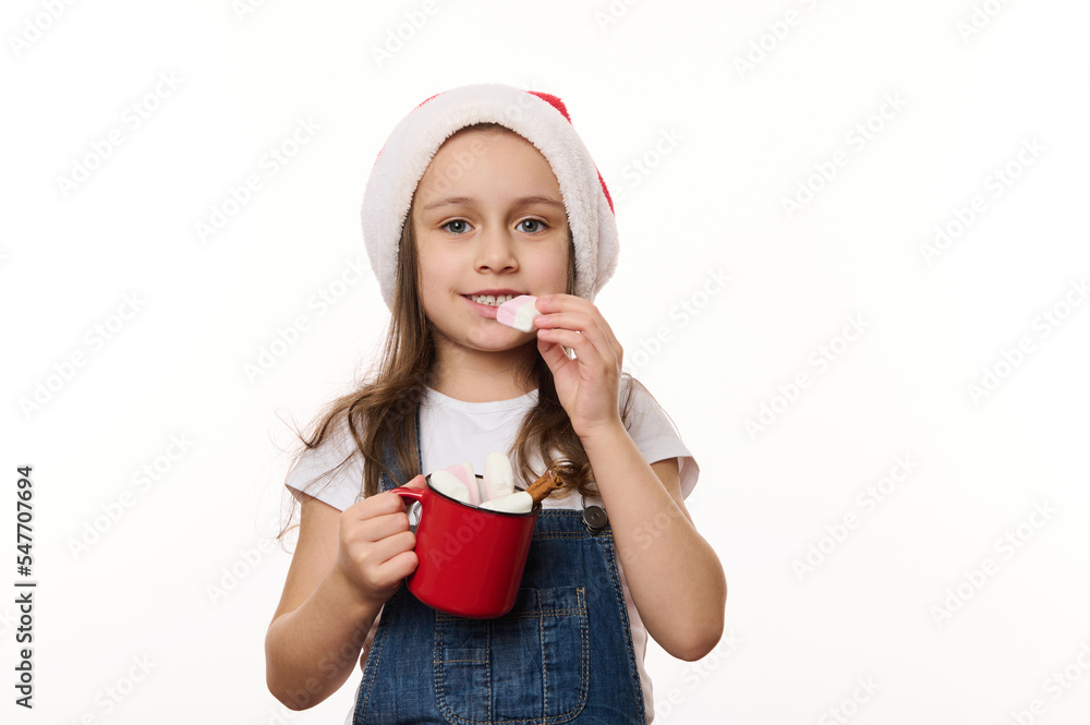 Isolated portrait on white background of lovely little child girl, in Santa hat, holding a red cup of hot chocolate and eating marshmallows. Magic atmosphere of upcoming Christmas, winter holidays. Isolated portrait on white background of lovely little child girl, in Santa hat, holding a red cup of hot chocolate and eating marshmallows. Magic atmosphere of upcoming Christmas, winter holidays.