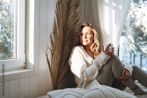 Young woman with blonde curly hair in white cardigan with cup of cocoa in hands looking at window with winter landscape at home photo