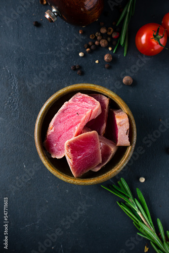 Pieces of tuna in a bowl with rosemary and spices on a dark background.