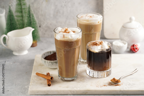 Three tall glasses with warm coffee drink with cinnamon, whipped milk foam and caramel on a marble board and christmas ornaments and decoration
