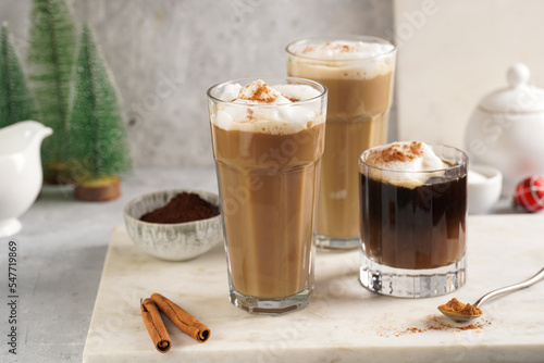 Three tall glasses with warm coffee drink with cinnamon, whipped milk foam and caramel on a marble board and christmas ornaments and decoration