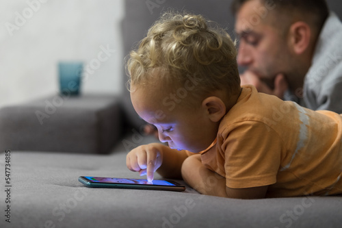 Toddlers and gadgets. Little boy using smartphone while lying on sofa with father at home, kid child playing mobile game on cellphone. Gadget addiction in children. Selective focus