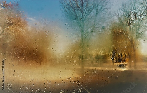 Feuchte beschlagene Glasscheibe mit Regentropfenmuster vor Schneelandschaft mit B  umen und Himmel bei Sonne am Mittag im Winter