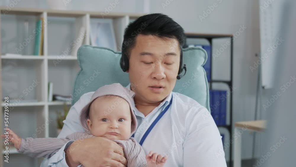 Multitasking father sitting at computer at office and talking care of baby. Footage of cute little boy and daddy. Single man in headset holding son in arms, talking. Video call Multitasking father sitting at computer at office and talking care of baby. Footage of cute little boy and daddy. Single man in headset holding son in arms, talking. Video call