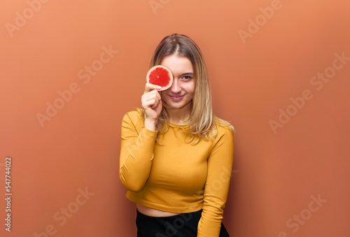 Young attractive girl covering her eye with slice of grapefruit, smiling, looking at camera, in yellow blouse and black bottom on orange background with copy space.
