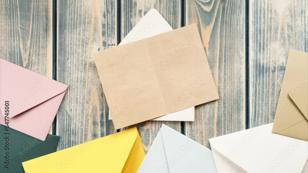 Looped stop motion video of white opening envelope on gray wooden background. Multicolored envelopes fall from above, kraft paper card is taken out of envelope and opened. Copy space. Top view