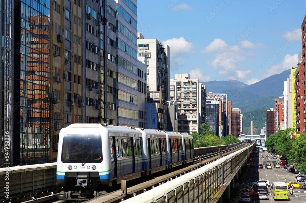Naklejka premium View of a train traveling on elevated rails of Wenhu Line in Taipei Metro System by office towers under blue clear sky ~View of MRT railways in Taipei, capital city of Taiwan, on a beautiful sunny day